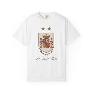 Può includere: Una t-shirt bianca a maniche corte con una grafica centrale dello stemma spagnolo nei toni dell'oro e del rosso, sormontato da due stelle dorate. Sotto lo scudo, il testo La Furia Roja è stampato in un carattere corsivo nero.