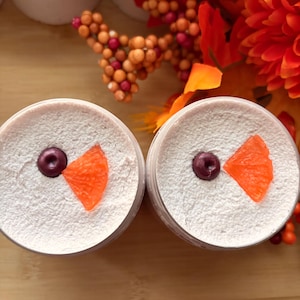 May include: Two round containers filled with a textured, off-white cream substance. Each container is topped with a single dark purple circular accent and a bright orange triangular piece, resembling a stylized face on the surface.