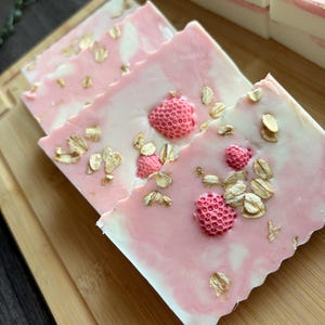 May include: Rectangular bars of soap featuring a marbled white and pink base. The surface of each bar is embedded with small, textured pink strawberry-shaped accents and scattered light brown oat flakes throughout the top layer.
