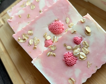 May include: Rectangular bars of soap featuring a marbled white and pink base. The surface of each bar is embedded with small, textured pink strawberry-shaped accents and scattered light brown oat flakes throughout the top layer.