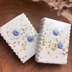 May include: Two square, white soaps with a scalloped edge, each with three light blue embellishments. The soaps are speckled with dried herbs and spices. The soaps are displayed on a wooden surface.