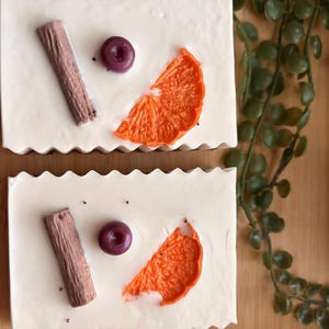 May include: Two rectangular bars of white soap with a scalloped top edge. Each bar is decorated with a brown cinnamon stick, a small purple berry, and a bright orange citrus slice embedded on the smooth, flat surface of the soap.