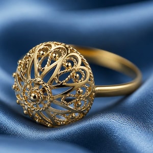 May include: A gold filigree ring with a round, openwork design. The ring features intricate patterns and small, decorative beads. The band is smooth and polished. The ring is set against a blue background.