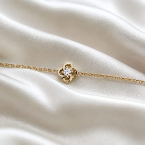 May include: A delicate gold chain bracelet featuring a central four-leaf clover motif. The clover design is crafted from polished gold metal and holds a single, clear, round-cut gemstone at its center, secured by four small prongs.