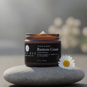 Könnte beinhalten: Ein braunes Glas mit "Restore Cream" und einem weißen Klecks oben, auf einem grauen Stein. Das Etikett lautet "BIS BEE" und "HAND & BODY". Eine weiße Gänseblümchen mit gelber Mitte liegt neben dem Glas. Das Glas enthält 60 ml.