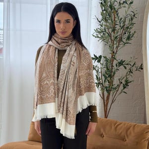 May include: A beige and white paisley patterned scarf with a fringed edge. The scarf is draped over a person's shoulders and chest. The scarf has a detailed design with a cream-coloured border.