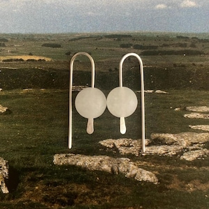 Puede incluir: Un par de pendientes colgantes plateados. Cada pendiente presenta una esfera blanca esmerilada suspendida de un alambre plateado curvado. La esfera está conectada a una pequeña gota plateada alargada. El fondo muestra colinas verdes y marrones.