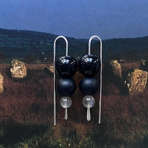 Puede incluir: Pendientes colgantes con un diseño de cuentas azul oscuro. Cada pendiente presenta un gancho plateado, dos cuentas esféricas azul oscuro, una cuenta azul oscuro más pequeña y una cuenta transparente en forma de lágrima. El fondo muestra un paisaje con cielo azul.