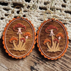 May include: A pair of wooden earrings featuring a mushroom design. The earrings have a brown background with orange and red mushrooms. The earrings are oval-shaped with a scalloped orange border and gold-colored ear wires.