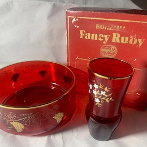 May include: A ruby red glass bowl and a matching vase with gold rim accents, accompanied by a red cardboard box labelled Bohemia Fancy Ruby. The bowl features gold etched grape vine patterns, while the vase displays a white floral design.