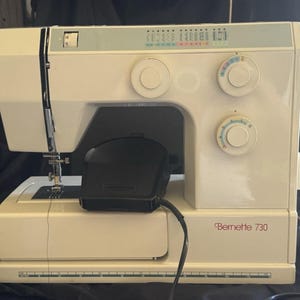 Puede incluir: Una máquina de coser Bernette 730 blanca con una pantalla de selección de puntadas en la parte superior, dos diales de control circulares en el frente, una aguja y un pedal negro sobre la base. Una regla está impresa en el borde inferior.