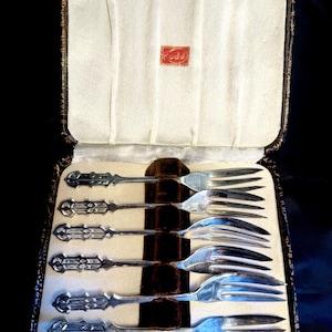 May include: A set of six silver-toned metal forks with ornate, geometric handles, arranged horizontally in a rectangular, hinged presentation case. The interior of the case is lined with cream-colored fabric and features a red label with the text Rodd.