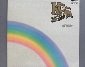 May include: A square vinyl record cover featuring a white background with a soft-focus rainbow arching from the bottom left. The top right displays the gold and black text KC AND THE SUNSHINE BAND, the RCA Victor logo, and the text INCLUDES THE HIT SHAKE, SHAKE, SHAKE SHAKE YOUR BOOTY.