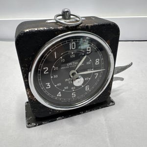 Puede incluir: Un temporizador industrial vintage de Smiths con carcasa de metal negro desgastado y esfera circular. La esfera muestra números blancos del 1 al 10, marcas de minutos interiores hasta 120 y manecillas plateadas. Un anillo de metal se sitúa en la parte superior.