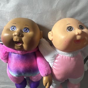May include: Two bald baby dolls with round faces and large eyes. The left doll has dark skin, purple eyes, and wears a pink and purple gradient plush outfit. The right doll has light skin, brown eyes, and wears a white and pink plush outfit.