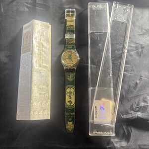 Puede incluir: Un reloj de pulsera Swatch Quartz con esfera dorada y el logotipo de los Juegos Olímpicos de Atlanta 1996. La correa verde oscuro muestra anillos olímpicos dorados y nombres de ciudades. Incluye una caja dorada y un estuche transparente.
