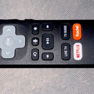Op de afbeelding: Een zwarte afstandsbediening met een centraal grijs navigatiepad met een OK-knop, een rode aan-uitknop en speciale knoppen voor Netflix, Foxtel en Live TV. Een klein paars label aan de zijkant toont de tekst Roku Powered in witte letters.