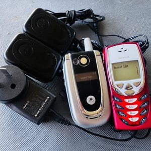 May include: A red Nokia mobile phone with a silver keypad, a silver flip phone with a black screen displaying Insert SIM, two black rectangular power adapters, and a black Nokia wall charger with a two-prong plug and attached power cord.