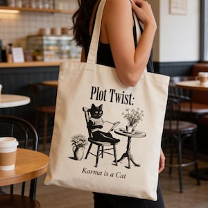 May include: A cream-coloured tote bag featuring the text "Plot Twist: Karma is a Cat" and a black and white illustration of a cat reading. The cat is seated at a small table with flowers. The bag has two long handles.