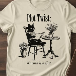 May include: A cream-coloured t-shirt with a black illustration of a cat reading a book at a table. The text above reads "Plot Twist:" and below "Karma is a Cat."