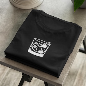 Puede incluir: Una camiseta negra con un estampado gráfico blanco de una copa de cóctel que contiene cubitos de hielo y dos cerezas. El diseño está centrado en la zona del pecho de la prenda doblada, mostrando una sencilla ilustración lineal de una bebida.