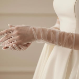 Könnte beinhalten: Elegante, transparente weiße Handschuhe mit zartem, fließendem Design. Die Handschuhe reichen bis zum Ellbogen und werden mit einem weißen Kleid kombiniert, wodurch ein anmutiger Look entsteht. Ideal für Hochzeiten oder formelle Anlässe.
