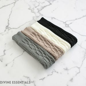 May include: A collection of five cable-knit leg warmers in various colors: gray, tan, cream, and black. The leg warmers are arranged diagonally on a marble surface. The text "DIVINE ESSENTIALS" is at the bottom left.
