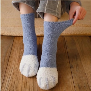 May include: A pair of cozy, light blue socks with white paw-shaped designs on the toes. The socks are made of a soft, fuzzy material, perfect for lounging. The image shows the socks being stretched.