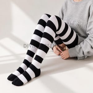 May include: A pair of black and white striped thigh-high socks. The socks have bold stripes and are worn by a person sitting on a white surface. The socks are made of a soft material, ideal for adding a playful element to any outfit.
