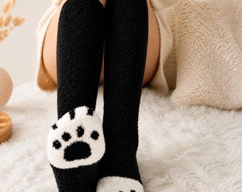 May include: Black knee-high socks with a white paw print design on the foot. The socks are made of a soft, fuzzy material. The text "DIVINE ESSENTIALS" is at the top of the image.