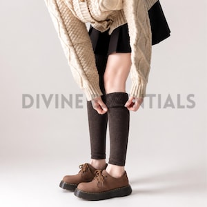 May include: A pair of dark brown knit leg warmers with a ribbed texture at the top, worn with a beige cable-knit sweater, a black skirt, and brown lace-up shoes with thick soles. The text DIVINE ESSENTIALS is visible in the center of the image.