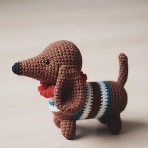 Pode incluir: Figura de cão dachshund de crochê em tons de castanho, com nariz preto e cachecol vermelho. O cão tem um suéter listrado em castanho, branco e verde-azulado. Um item feito à mão, charmoso e decorativo.