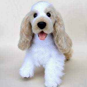 Pode incluir: Um brinquedo de pelúcia de cachorro Cocker Spaniel com corpo branco, orelhas castanhas e nariz preto. A boca do cão está aberta, revelando uma língua rosa. O brinquedo foi projetado para parecer realista e é feito de material macio e fofo.