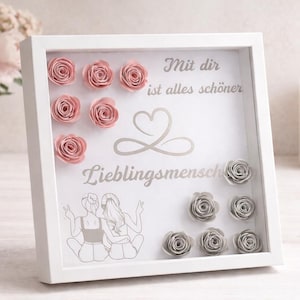 Könnte beinhalten: Ein weißer Schattenrahmen mit Papierrosen. Die Box zeigt rosa und graue Papierrosen, den deutschen Text "Mit dir ist alles schöner" und "Lieblingsmensch" sowie eine Zeichnung von zwei Personen. Eine Kerze und Blumen sind im Hintergrund.