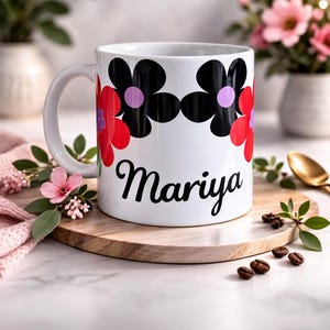 Op de afbeelding: Witte keramische mok met een bloemenontwerp in rood, zwart en paars. De mok heeft de naam "Mariya" in een zwart scriptlettertype. De mok staat op een houten plank met koffiebonen en groen.