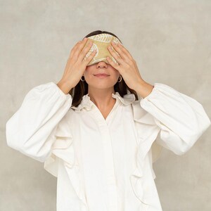 Puede incluir: Una máscara para dormir beige con un estampado de hojas cubre los ojos. La máscara se sujeta con las manos de una persona que lleva una blusa blanca y vaqueros azules. La máscara para dormir está diseñada para bloquear la luz.