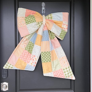 May include: A decorative fabric bow in a patchwork design, hanging on a black door. The bow is made of squares in pastel shades of pink, green, yellow, and blue, with floral and dotted patterns. A charming home decor item.