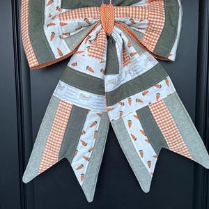 May include: A large, decorative bow made of fabric, featuring a patchwork design with orange, green, and white patterns. The bow includes carrot and bunny prints, and gingham patterns. It is designed for seasonal or holiday decor.