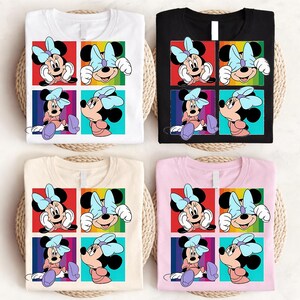 May include: Four t-shirts in white, black, beige, and pink, each featuring a colorful Minnie Mouse graphic. The graphic shows Minnie in four different poses within a grid of colorful squares. The shirts are displayed on a woven surface.