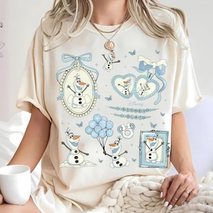 May include: A cream-coloured t-shirt with multiple illustrations of Olaf from Disney's Frozen. The designs include Olaf in various poses, surrounded by blue accents, bows, and butterflies. The text "Disney Olaf" is also present.
