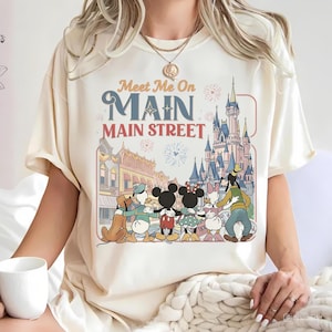 Puede incluir: Camiseta color crema con un gráfico colorido de personajes de Disney, incluyendo Mickey y Minnie Mouse, Donald Duck y Goofy, con el texto "Meet Me On Main Street". El diseño incluye un castillo y edificios de Main Street.