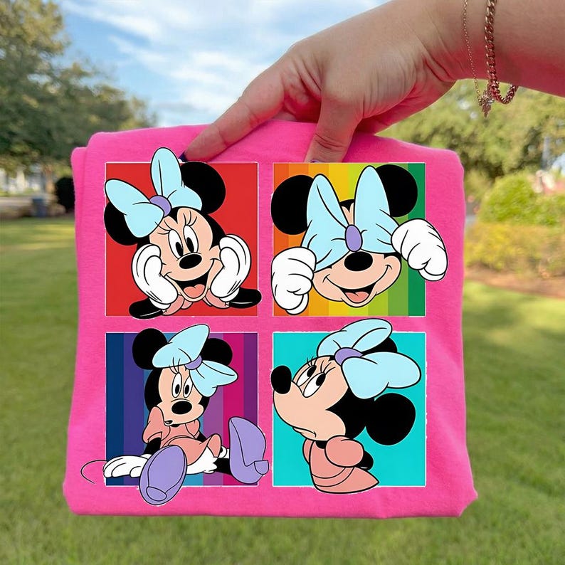 May include: A pink t-shirt featuring four panels of Minnie Mouse illustrations. Each panel showcases Minnie in different poses, wearing her signature light blue bow. The background colors vary, including red, rainbow stripes, blue, and turquoise.