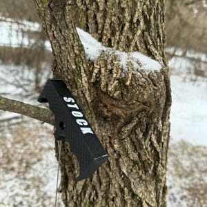 May include: A black textured grip with the word "STOCK" in white lettering is attached to a tree. The grip has a curved design and is made of a durable material. The tree bark is brown and textured, with a small patch of snow.