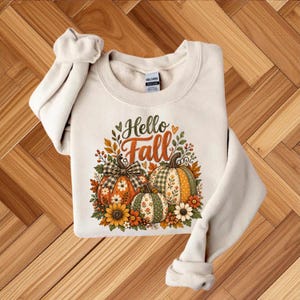May include: A cream-coloured crewneck sweatshirt featuring a graphic print of three pumpkins with various patterns, including plaid and floral designs. The text "Hello Fall" is printed in brown and orange above the pumpkins, surrounded by leaves.