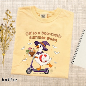 May include: A butter yellow t-shirt featuring a graphic of a white duck wearing a purple witch hat and scarf, riding a purple scooter while holding a coconut drink and a beach ball, with the text Off to a boo-tastic summer ween and bats.