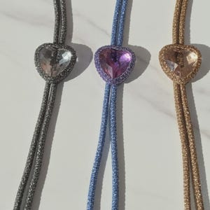 Puede incluir: Tres corbatas bolo en forma de corazón. Una es plateada con un corazón transparente, otra es azul con un corazón morado y la última es dorada con un corazón melocotón. Cada corbata tiene un cordón brillante y puntas plateadas.