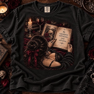 Może przedstawiać: Ciemnoszary t-shirt z nadrukiem graficznym przedstawiającym osobę z ciemnymi włosami związanymi czerwoną wstążką, czytającą otwartą książkę. Książka zawiera tekst 'My redemption will become your salvation' oraz wizerunek czaszki, świecy i wina.