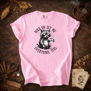 May include: A light pink t-shirt featuring a black and white illustration of a raccoon holding a mug. The text on the shirt reads "Hold on let me overthink this". The mug says "Soft Menace".