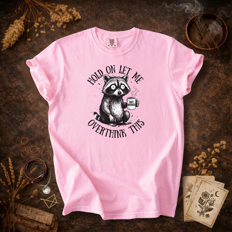 May include: A light pink t-shirt featuring a black and white illustration of a raccoon holding a mug. The text on the shirt reads "Hold on let me overthink this". The mug says "Soft Menace".