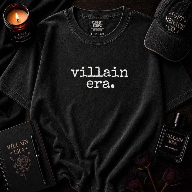 May include: Black t-shirt with the text "villain era." A black baseball cap with "+SOFT+ MENACE +CO.+" is visible. A black notebook with "VILLAIN + ERA+" and a rose graphic is also present. A candle and perfume bottle are also in the image.